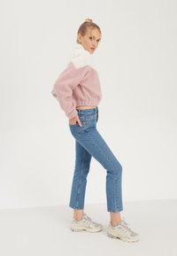 Sweater en polaire colorblock rose et blanc, coupe cropped ; associé à un jean droit bleu clair et des baskets grises. Fond blanc.