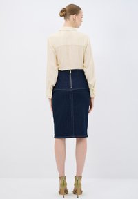 Camisa de manga larga beige con puños de botón, combinada con una falda lápiz de mezclilla oscura con cremallera trasera y costuras en contraste. Tacones oliva.