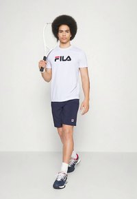 Valge Fila T-särk punase ja sinise logoga, tumesinised lühikesed püksid ja hallid tossud. Hoides tennise reketit, seisab neutraalse tausta ees.