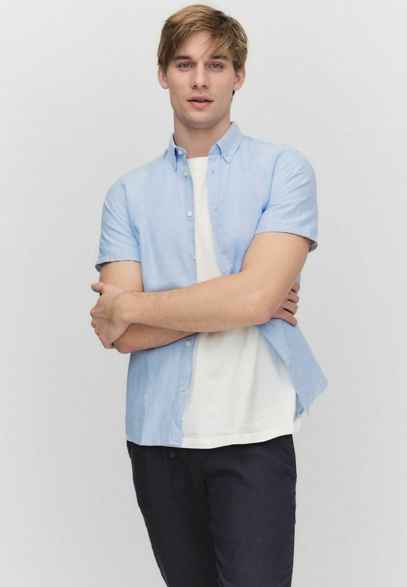 Lichtblauw kortom mouw overhemd over een wit t-shirt, gecombineerd met donkere broeken; casual fit met zichtbare knopen en een relaxte kraag.