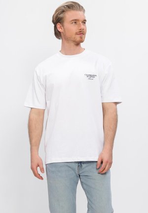 Blonder Mann trägt ein weißes T-Shirt mit der Aufschrift "Celebration of Speed Lakewood" und hellblaue Jeans, steht vor einem weißen Hintergrund.