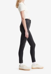 Femme portant un haut court à manches courtes blanc, un jean skinny noir taille haute, des chaussettes blanches et des baskets crème à semelles gomme, debout de profil.