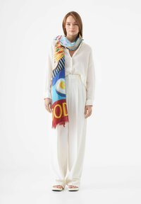 Écharpe colorée à motifs dans des tons de rouge, bleu et jaune, portée sur une chemise en lin blanche et un pantalon large blanc. Tissu doux et léger.