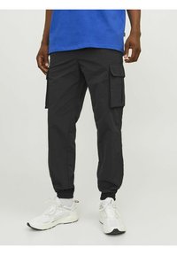 Sorte cargo bukser med elastisk talje, der har flere sidelommer og et tapered ben-design. Matchet med hvide træningssko.