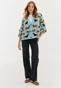 Svetle modrý pletený cardigan s motivem psa, se zapínáním na knoflíky, spadlými rameny a žebrovanými manžetami, v kombinaci s tmavými džíny.