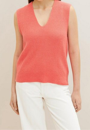 Vrouw draagt een mouwloze koraalrode gebreide top met V-hals, gecombineerd met een hoge taille witte broek tegen een neutrale achtergrond.