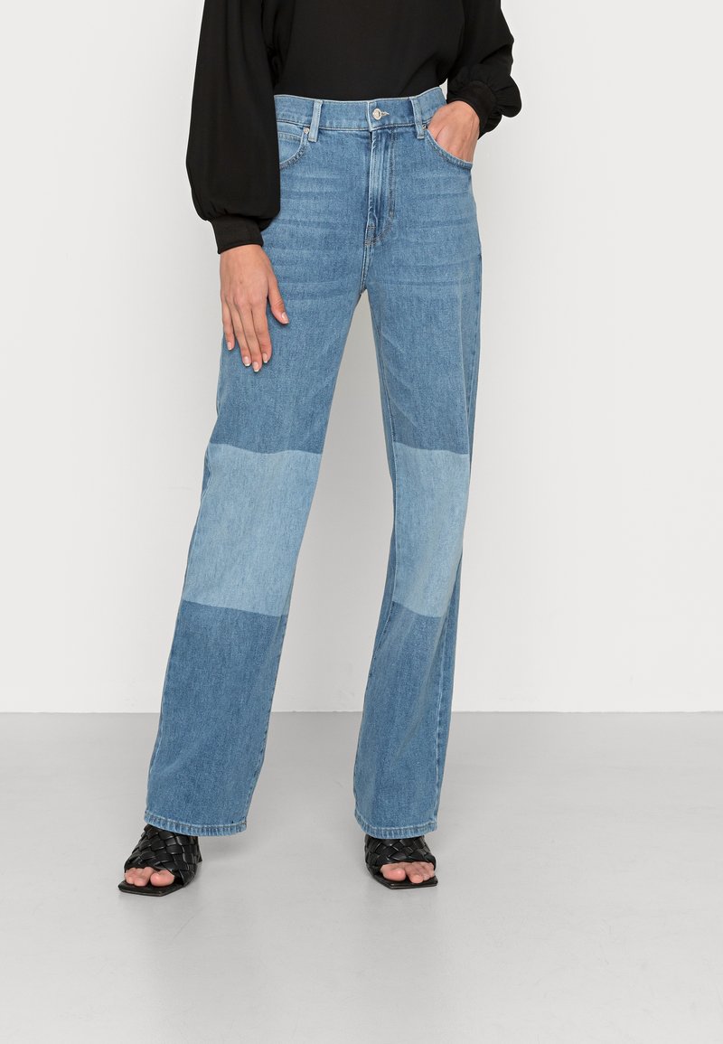 Jeans de ganga de perna larga em azul claro, com design em patchwork, bolsos frontais e uma cintura alta. Estilizados com sandálias pretas de fibra trançada e uma blusa preta.