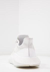 Valkoinen urheilukenkä, jossa on teksturoitu neulottu päällinen, saumaton muotoilu, pehmustettu nilkkakappale ja kuvioitu kumipohja. Minimalistinen ja moderni ilme.