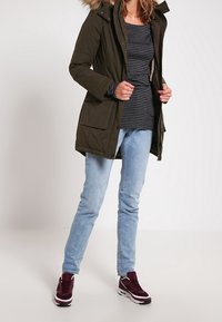 Woman wearing dark green parka with fur hood, striped long-sleeve shirt, light blue jeans, and maroon sneakers standing against plain background.