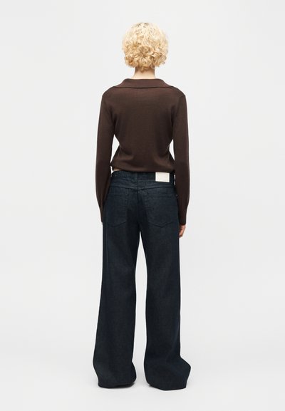 Άτομο με κοντά σγουρά ξανθά μαλλιά φοράει καφέ μπλούζα με μακριά μανίκια και άνετο σκουρομπλε τζιν, στέκεται στραμμένο σε μια απλή ανοιχτόχρωμη επιφάνεια.