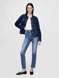 Jaqueta de denim escuro com fecho de botão, dois bolsos no peito e um design ligeiramente curto, combinada com jeans azul claro e sapatilhas pretas.
