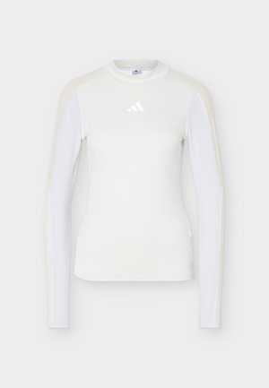 Dametopp med lange ermer, hvit og kroppsnær, med rund hals og Adidas-logo på brystet, med subtile beige skulderstriper.
