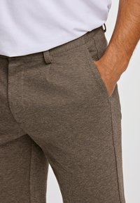Bruine, gestructureerde broeken met een subtiel patroon, voorzien van zijzakken en een gladde tailleband, gecombineerd met een wit shirt.