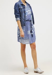 Veste en denim sur une robe bleue à motifs avec une taille nouée. Chaussures oxford en argent. La robe présente divers motifs géométriques et floraux.