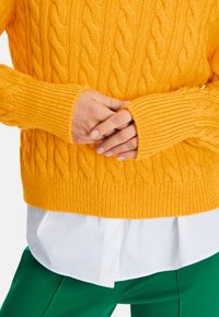 Helles gelbes Kabelstrickoberteil mit gerippten Bündchen und Saum, über einem weißen Hemd getragen, kombiniert mit grünen Hosen.