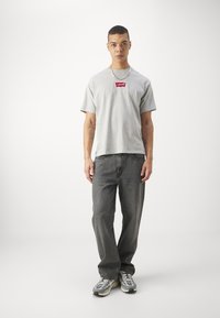 Grå kortærmet T-shirt med rødt Levi's-logo, parret med mørkegrå jeans og grå sneakers. Har en afslappet pasform og rund hals.