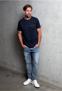 Námořnické tričko s vyraženým logem, světle modré džíny a bílé tenisky, stojící proti betonové zdi. Jednoduchý, neformální outfit.