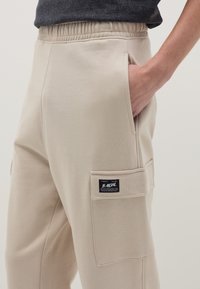 Beige Jogginghose mit elastischem Bund, seitlichen Taschen und einer aufgenähten Tasche am Oberschenkel mit einem schwarzen Label-Logo. Weicher Stoff.