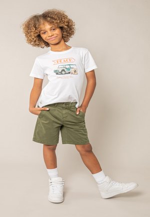 Kind met krullend haar, gekleed in een wit "Beach" t-shirt, olijfgroene korte broek, witte sokken en witte sneakers, staand met handen in de zakken en één been uitgestrekt.