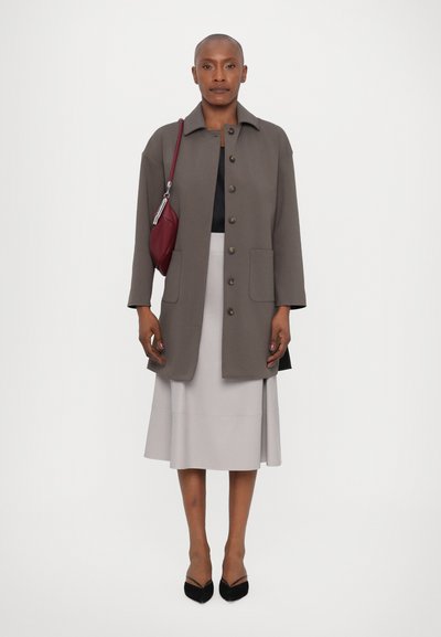 Grau gestepptes Oberbekleidungsstück mit Kragen, Fronttaschen und kontrastierenden Knöpfen, kombiniert mit einem hellgrauen Rock und schwarzen spitzen Schuhen.