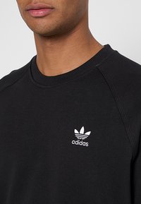 Svart sweatshirt med rund halsringning och ribbade ärmslut. Har en vit Adidas-logotyp på vänster bröstområde. Slät textur.