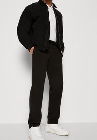 Svart zip-up jacka med krage, vit polo skjorta under, och svarta byxor. Modellen bär vita sneakers och står mot en ljus bakgrund.