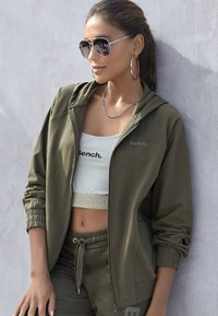 Oliwkowy strój sportowy, w skład którego wchodzi zapinana na zamek bluza z kapturem, krótka bluzka oraz dresy z elastycznym pasem, z logo "Bench." i metalowymi akcentami.