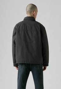 Mand med barberet hoved iført sort jakke og blå jeans, vendt væk mod en simpel lys grå baggrund.
