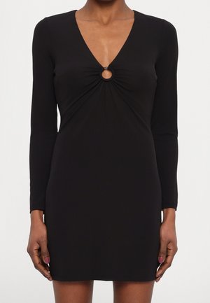 Robe mini noire à manches longues avec décolleté en V et détail d'ouverture circulaire au niveau de la poitrine, portée par une personne debout, les bras détendus.