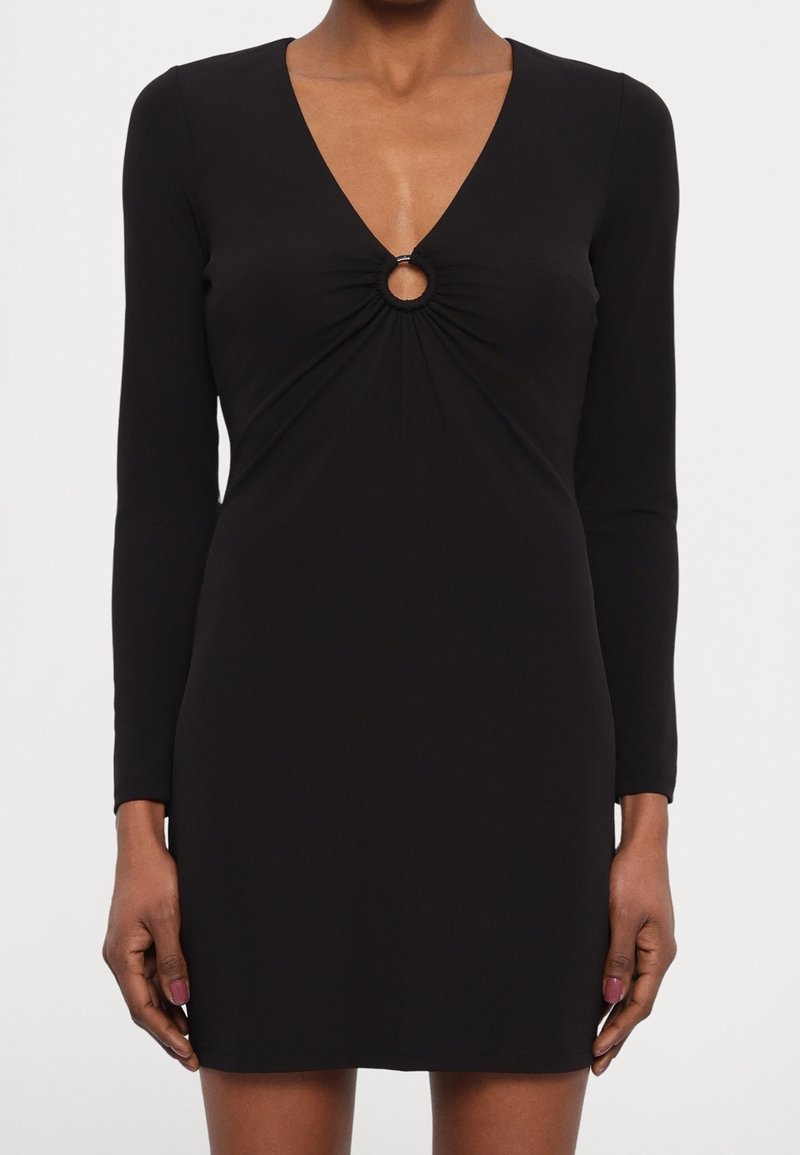 Robe mini noire à manches longues avec décolleté en V et détail d'ouverture circulaire au niveau de la poitrine, portée par une personne debout, les bras détendus.
