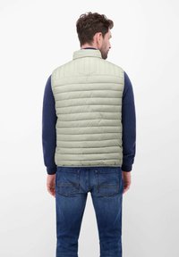 Jasnozielona, wyściełana kamizelka z wysokim kołnierzem i pionowym pikowaniem, noszona na granatowej teksturowanej koszuli z długim rękawem, zestawiona z dżinsami.