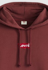 Bordo dukserica s kapuljačom na vezanje, s bijelim i crvenim izvezenim Levi's logom. Tkanina je teksturirana i pruža opuštenu fit.