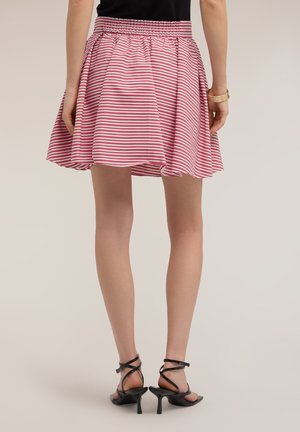 Vrouw draagt een korte rood-wit gestreepte rok met een zwart topje en zwarte sandalen met hoge hakken, staand tegen een effen achtergrond.