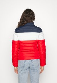 Kvinna med långt lockigt hår iklädd en blå, vit och röd pufferjacka och ljusblå jeans, stående vänd bort från kameran mot en enfärgad bakgrund.