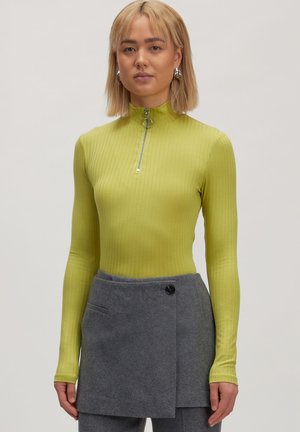 Rippstrick-Longsleeve-Oberteil in leuchtendem Limettengrün mit hohem Kragen und halbem Reißverschluss, kombiniert mit einem grauen, geknöpften, asymmetrischen Rock.