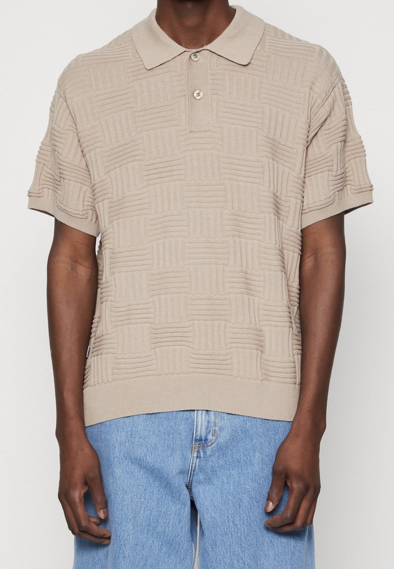 Homme portant un polo beige texturé à manches courtes et un jean en denim bleu clair sur fond blanc.