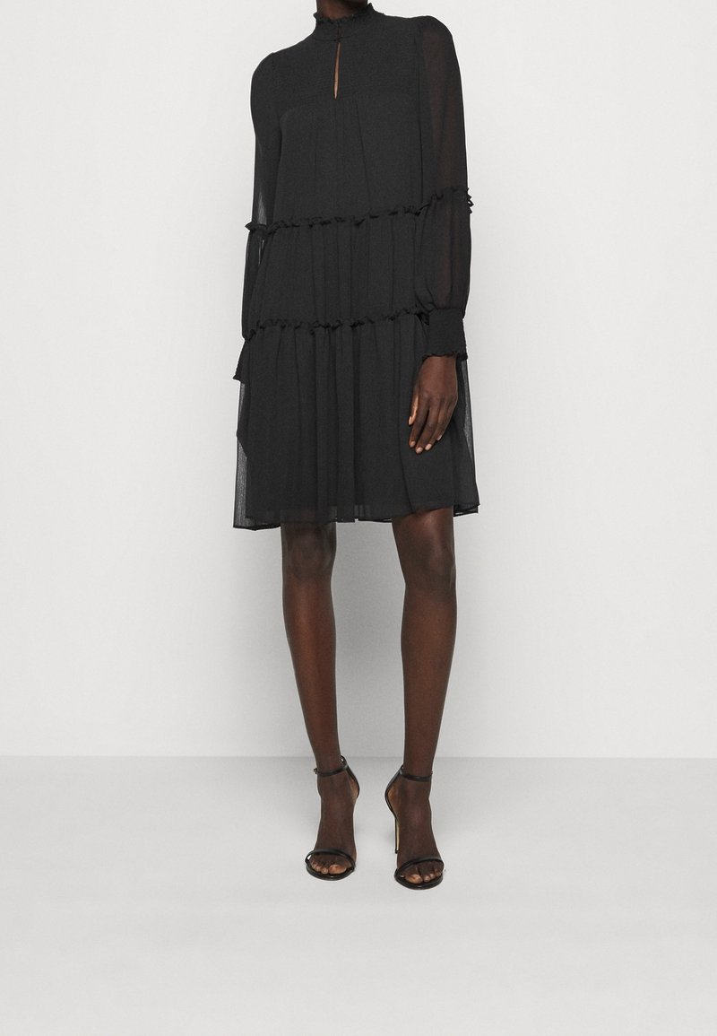 Femme portant une robe noire transparente à volants, longueur genoux, manches longues, et des sandales noires à talons hauts avec une bride à la cheville.