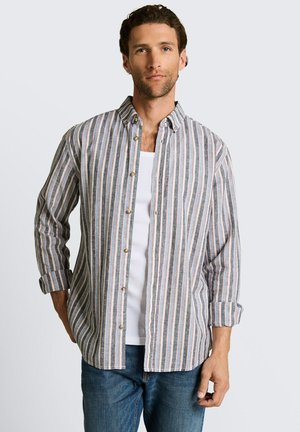 Uomo che indossa una camicia a righe a maniche lunghe sbottonata sopra una canottiera bianca e jeans blu, in piedi davanti a uno sfondo chiaro e semplice.