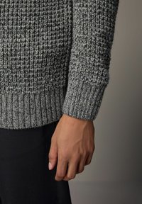 Grå strikket sweater med et tekstureret mønster, der har en ribbet kant og manchetter, sammen med mørke bukser, fremhævet af en hånd der hviler mod siden.