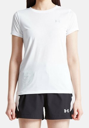 Žena v jednoduchém bílém sportovním tričku s krátkým rukávem a černých sportovních šortkách s bílým logem na jednobarevném pozadí.