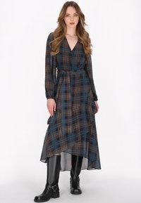 Lang geruit jurk van doorschijnende stof in donkerblauw, rood en wit, met een wikkelontwerp, lange mouwen en een strikdetail rondom de taille. Gecombineerd met zwarte kniehoge laarzen.
