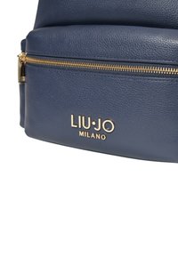 Zaino in pelle blu navy con hardware dorato, dotato di un compartimento con zip e logo del marchio in rilievo sulla parte anteriore. Dettagli con superficie testurizzata.