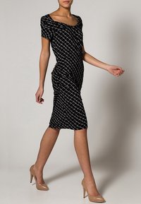 Svart figurnära klänning med ett vitt rutmönster, korta ärmar och rynkdetaljer, kombinerad med nakna högklackade skor. Slät tygstruktur.