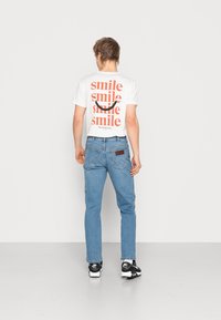 Vit T-shirt med upprepad text "smile" i orange, tillsammans med ljusblå jeans och svarta sneakers. Jeansens baksida har en etikett.
