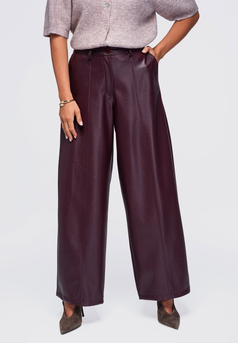 Femme portant un pantalon en cuir bordeaux foncé à jambes larges, un pull à manches courtes taupe et des talons pointus gris, debout avec une main dans la poche.