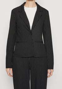 Blazer nero a righe sottili con bottone singolo, rever a punta e pantaloni abbinati a righe sottili indossati sopra una maglietta bianca, vista dalla vita in su.