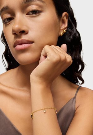 Pulsera de oro con gemas moradas y azules, que presenta un delicado diseño de cadena. Fondo con un tono de piel suave y una blusa de tela marrón oscuro.