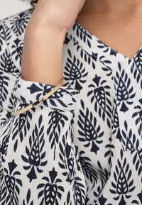 Käsi koukistettu lähellä kasvoja, päällä valkoinen paita mustilla lehtikuosisilla yksityiskohdilla ja kapealla kultaketjulla ranteessa.