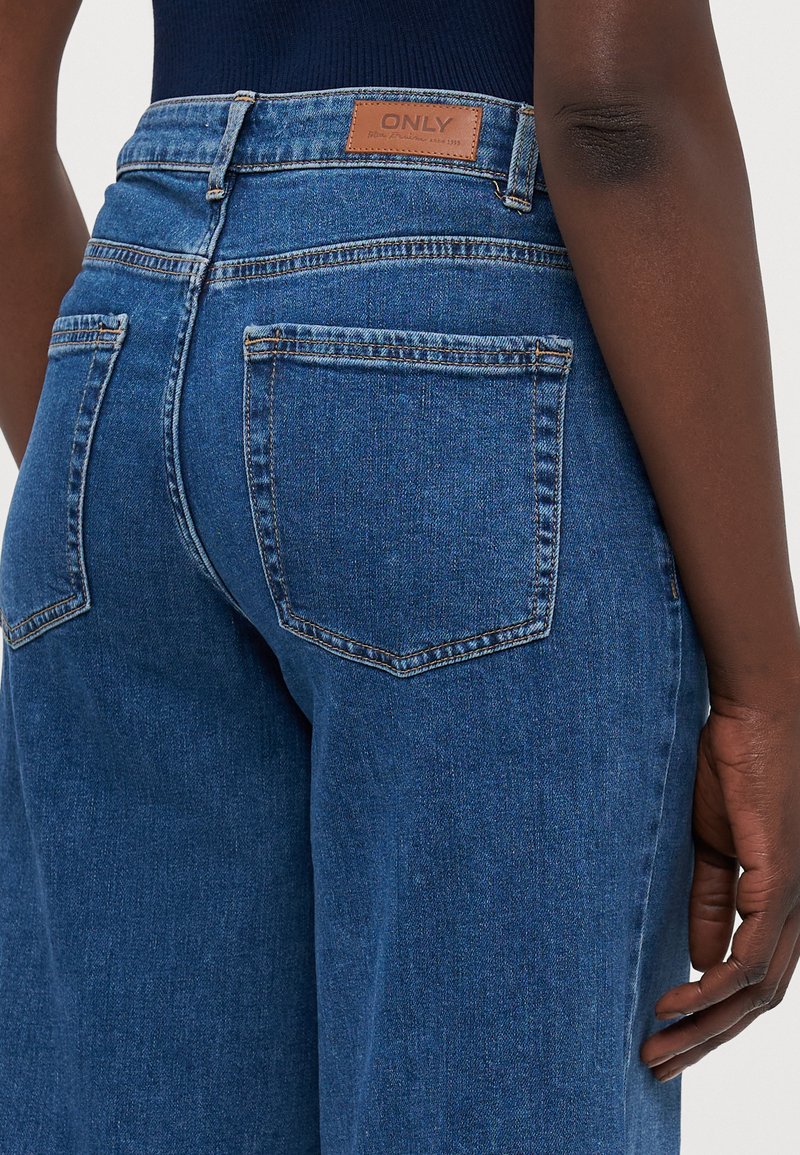 Jean en denim bleu à taille haute, avec une patch en cuir étiquetée "ONLY", et deux poches arrière avec des détails de couture. Texture lisse.