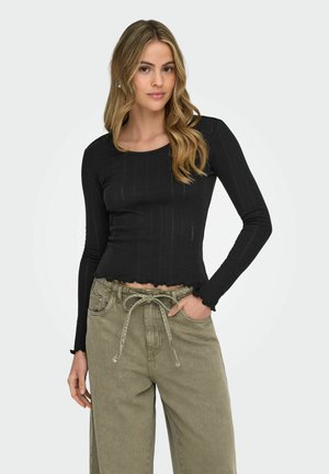 Woman with long blonde hair wearing a fitted black long-sleeve top and loose olive-green pants with a drawstring, posing with one hand in pocket.